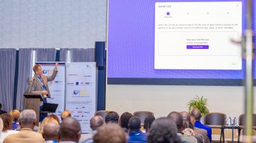 The image shows an event where a man is giving a presentation at a conference room with a large screen displaying information about the East African Humanitarian Summit. The attendees are seated, listening attentively as the speaker shares insights into the summit's objectives and the role of emerging technologies in supporting humanitarian efforts.