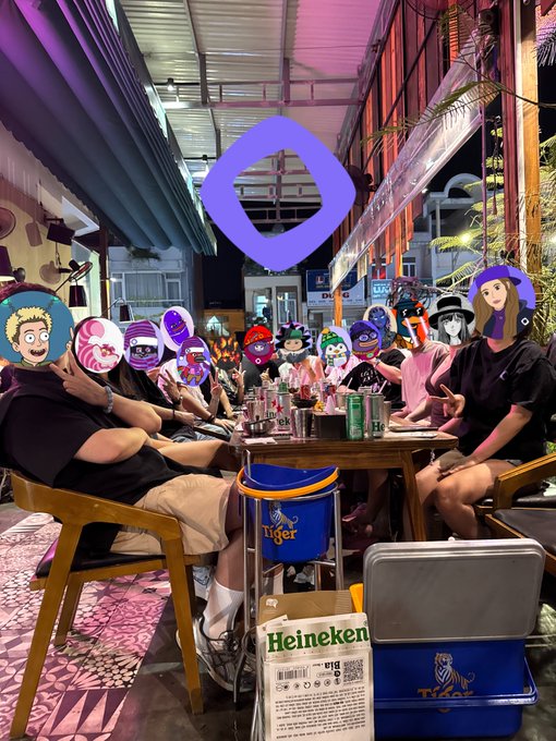 A lively group of people gather around a table at a bar with their faces covered by cartoon stickers, surrounded by Tiger and Heineken beer crates, while a large purple Monad logo hangs above them, capturing the casual "vietnads" meetup vibe referenced in the post.