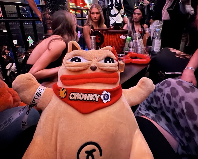 A tan plush cat toy with a red bandana reading 'CHONKY' is held up in the foreground of a nightclub or bar scene with people in the background.