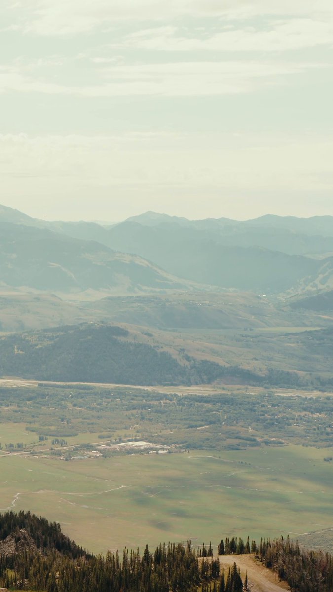 A sweeping aerial view captures the layered mountain ranges and vast green valley of Jackson Hole, Wyoming, serving as the scenic backdrop for Kraken's third annual participation in the SALT Conference and Wyoming Blockchain Symposium. The expansive landscape, with distant peaks fading into hazy skies above rolling hills and a small settlement below, mirrors the post's "big skies, bigger conversations" theme of high-level dialogue in crypto, finance, and policy.
