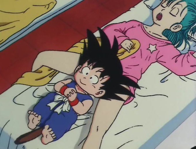 An animated scene showing a young Goku with spiky black hair lying on a bed next to a sleeping woman in pink pajamas with a star design, in a scene that appears to be from Dragon Ball.