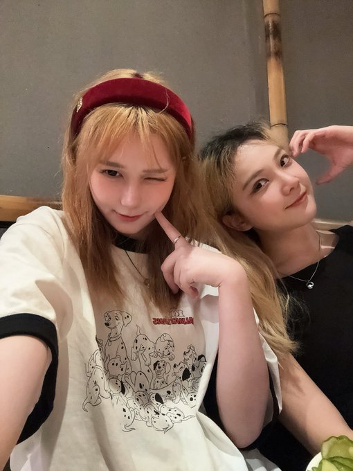 Two young women pose together for a selfie, one wearing a red velvet headband and a white t-shirt featuring Disney 101 Dalmatians characters, the other in a black top.