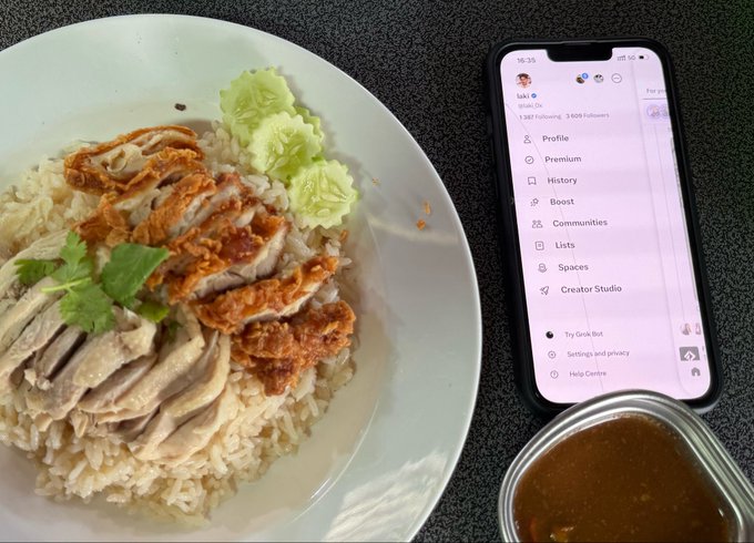 A top-down photograph of a meal consisting of rice topped with sliced chicken, fried egg, and cucumber garnish served on a white plate, alongside a phone displaying a social media profile menu and a small container of dipping sauce.
