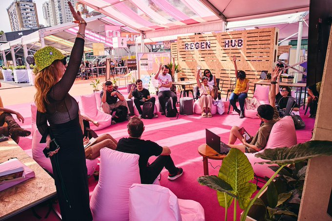 Attendees gather in a vibrant pink "REGEN HUB" community space at Devcon 8 in Mumbai, with one participant raising her hand while others sit on bean bags and engage in discussions. The colorful, casual setup reflects the post's message about Community Hubs being grassroots gathering spaces for shared ideas at the event.