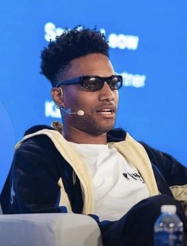 A young man with curly hair and black sunglasses speaks into a headset microphone while seated on a panel, appearing to discuss NFTs in relation to the announcement that "ANSEM SAYS NFTS ARE 'BACK'." Partial text reading "Sea..." and "son" is visible on the blue screen behind him.