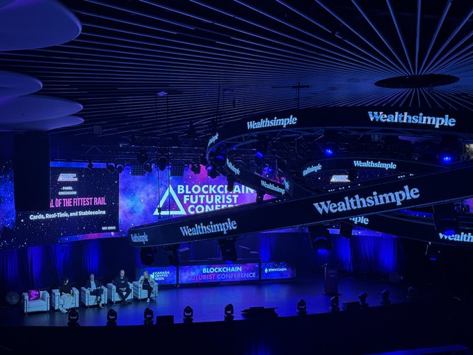 A panel discussion stage at the 'Blockchain Futurist Conference' featuring speakers seated on white sofas, with 'Wealthsimple' branding visible on overhead screens and banners, and a slide referencing Cardano and Stablecoins.
