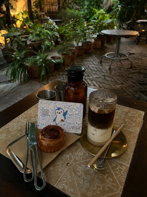 A coffee shop table featuring an egg tart, a layered iced coffee, a glass bottle, and a Visa credit card with a cute cartoon design, surrounded by plants.
