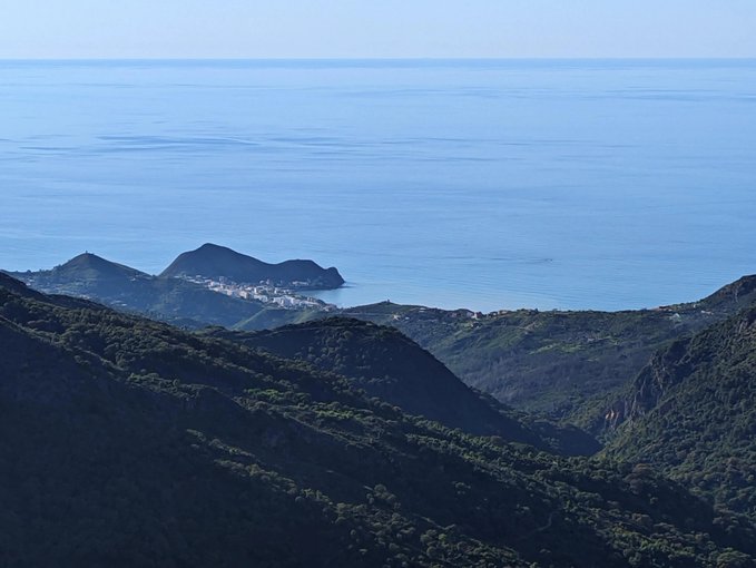 A scenic landscape photograph showing lush green mountainous terrain in the foreground overlooking a vast blue ocean, with a small coastal town visible nestled along the shoreline in the distance.
