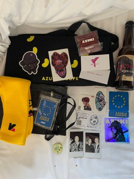 An assortment of merchandise spread on white sheets, including an Azuki beanie, yellow Vans cap, Azuki pins/stickers, a Polaroid photo, a black tote bag, an Azuki tumbler, and a bottle.