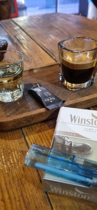 Two glasses of whiskey on a wooden table, alongside a pack of Winston Silver cigarettes, a tobacco pouch, and a blue lighter.