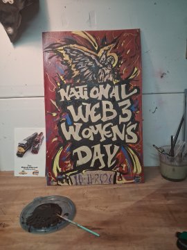 A wooden carved artwork displayed on a workbench celebrating 'NATIONAL WEB3 WOMEN'S DAY' on 11.11.2024, with paint and brushes nearby.