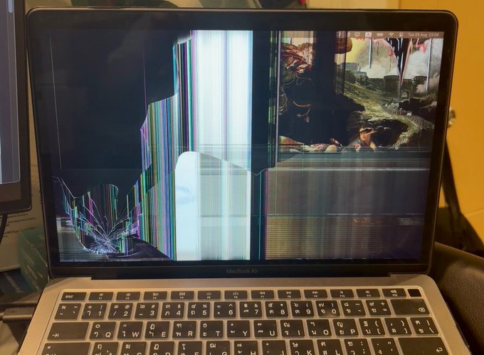 A MacBook Air with a severely cracked and glitching display sits open on a desk. The tweet "gm! should I mint this to pay for a new MacBook?" humorously suggests selling the broken laptop's glitched image as an NFT to fund a replacement.