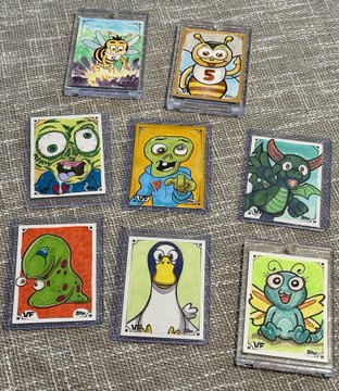 Eight hand-drawn/cartoon-style trading cards featuring cute monster characters including a bee, cat, green creatures, dragon, turtle, penguin, and frog, arranged on a textured grey surface.
