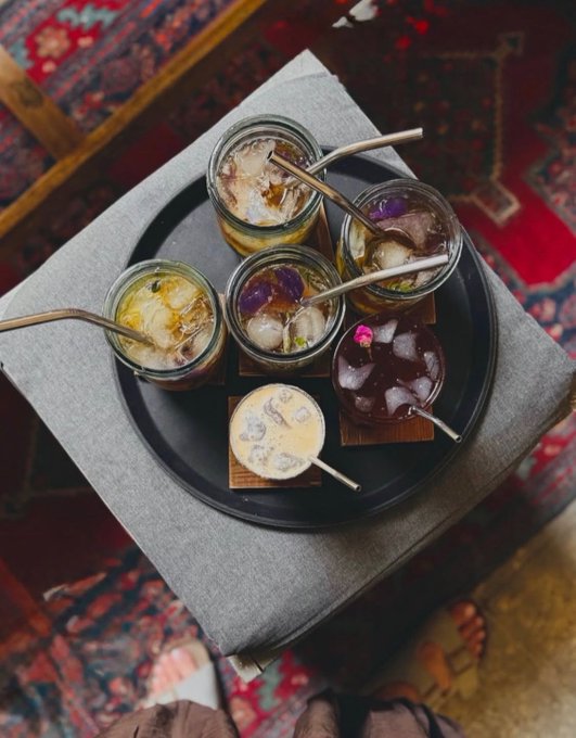 An overhead shot of a flight of various cocktails in small glasses served with metal straws, arranged on a black plate on a gray ottoman over a patterned red rug.