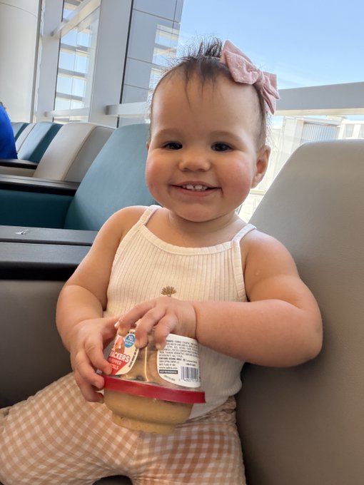 A smiling toddler wearing a pink bow and cream-colored tank top sits in a chair holding a Gerbers baby food container with the lid partially removed.