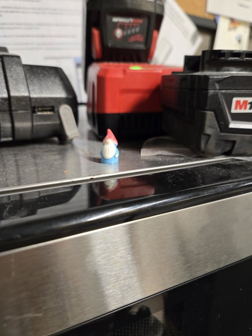 A small gnome figurine sits on a stainless steel surface in a workshop, with Milwaukee brand power tool batteries visible in the background.