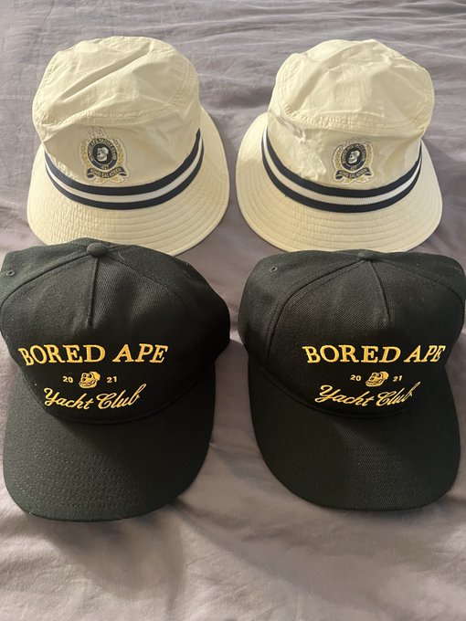 Four hats displayed on a surface: two white bucket hats with striped bands, and two black baseball caps with gold 'Bored Ape Yacht Club 2021' embroidery.