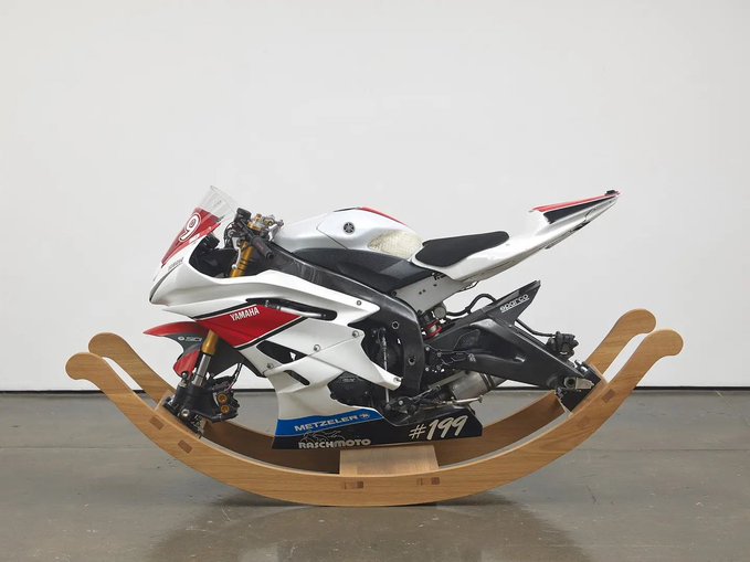 A white, red, and black Yamaha racing motorcycle marked with 'METZELER', 'RICHMOTO', and '#199' is mounted on a wooden rocking cradle against a plain gallery wall.
