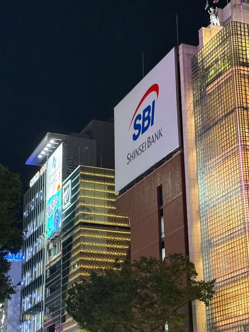 A nighttime cityscape shows a brightly lit building featuring a large billboard for "SBI Shinsei Bank" alongside illuminated structures and signage. The image illustrates the financial institution context, referencing SBI—the Japanese bank that has publicly expressed optimism about XRP, lending visual weight to the claim that "XRP will flip Bitcoin."