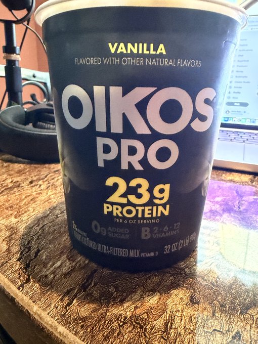 A container of Oikos Pro vanilla yogurt with 23g of protein, 0g added sugar, and 2.6g B-12 vitamins sits on a wooden desk, ready to be enjoyed for a protein-packed snack. The "Nom nom nom" caption suggests the delicious, satisfying taste of this high-protein treat.