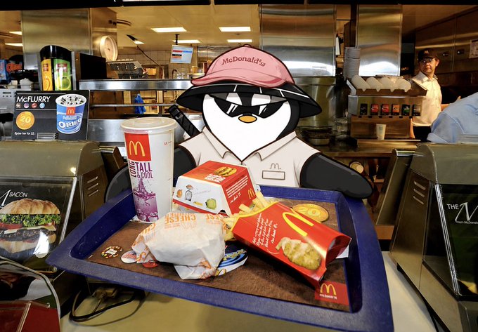 A cardboard cutout of a McDonald's employee wearing a "McDonald's" visor sits at a counter with a McDonald's meal tray containing a "McFlurry" cup, fries in a red carton, a burger wrapper, and a "Colonel's Cool" drink. In the background, employees work the kitchen, adding ironic humor to the post's boastful claim about working at McDonald's to earn "generational wealth."