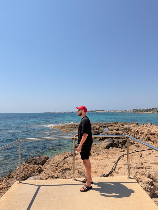 A man in a black outfit and red cap stands on a sunlit coastal platform, gazing out at the turquoise sea and rocky shoreline beneath a clear blue sky — a serene "good morning" scene capturing peaceful seaside vibes.