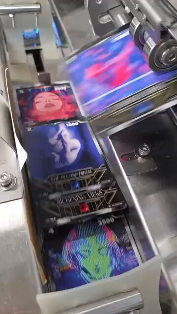 A close-up view of an industrial printing machine producing colorful album-style cards or stickers, with visible text including "THE WILL / NO FRIENDS," "BECOMING XENA," and numbers like "3500" and "4" printed on the glossy items. The "Gm" caption suggests a casual good morning greeting, paired with the imagery of a fresh production run starting up.