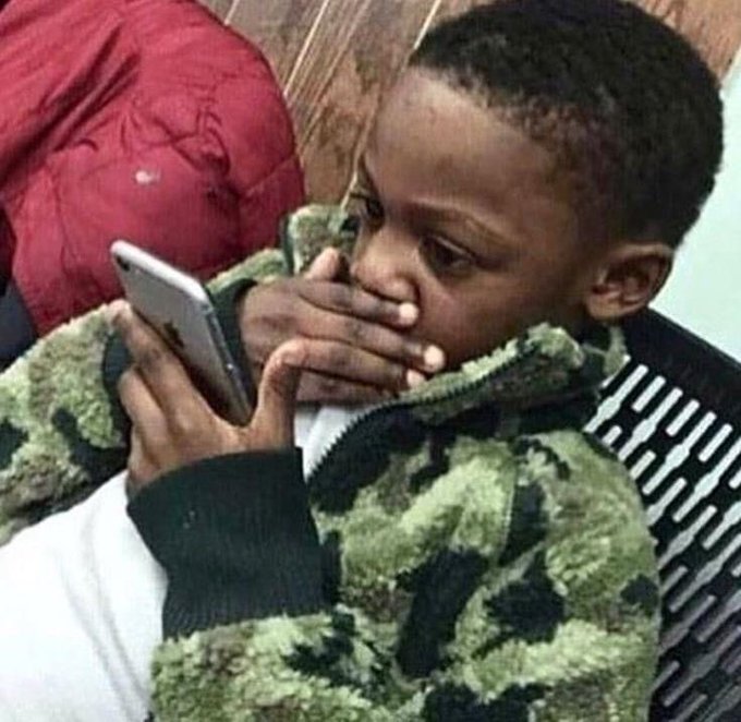 A young boy in a camouflage jacket sits with his hand over his mouth, appearing shocked or emotional as he looks at his phone.