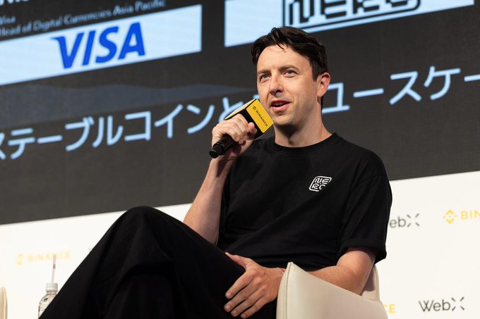 A man speaking into a yellow microphone at a panel discussion, with VISA and Binance logos visible in the background along with Japanese text 'ステーブルコイン' (stablecoin).