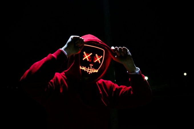 A person in a red hoodie with their hood up wears an illuminated mask featuring an X-eyed stitched smile, holding the hood closed against a dark background.