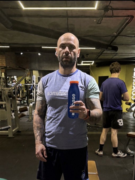 A bald, muscular man in a blue t-shirt stands in a gym holding a blue 'hase' brand water bottle, with another person in a purple shirt using equipment in the background.