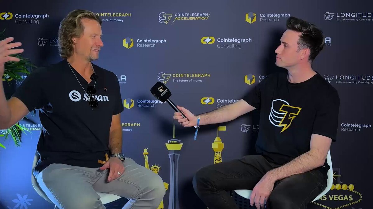 The image shows two men sitting next to each other, with a microphone between them, suggesting they are being interviewed or participating in an event. One of the men has a serious expression while the other appears more relaxed. The background features a blue wall adorned with various logos and text, including one that reads "Wall Street wants Bitcoin." This implies that there is some sort of discussion or event related to cryptocurrency or Wall Street's interest in it.