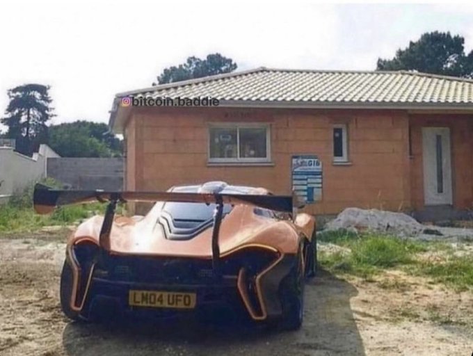 A copper-colored McLaren Senna with the license plate "LM04 UFO" and a large rear wing is parked on a dirt patch in front of a partially constructed house.