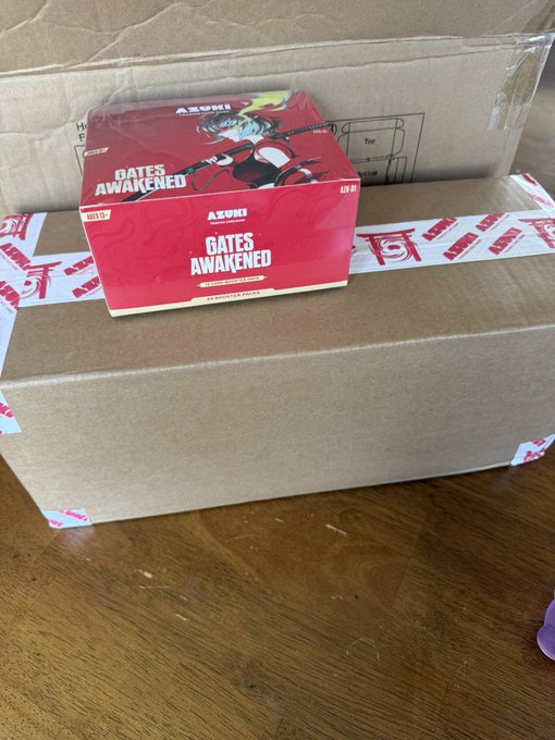 A photograph of a red 'AZUKI' 'GATES AWAKENED' trading card game booster box resting on top of a larger cardboard shipping box sealed with red and white tape.
