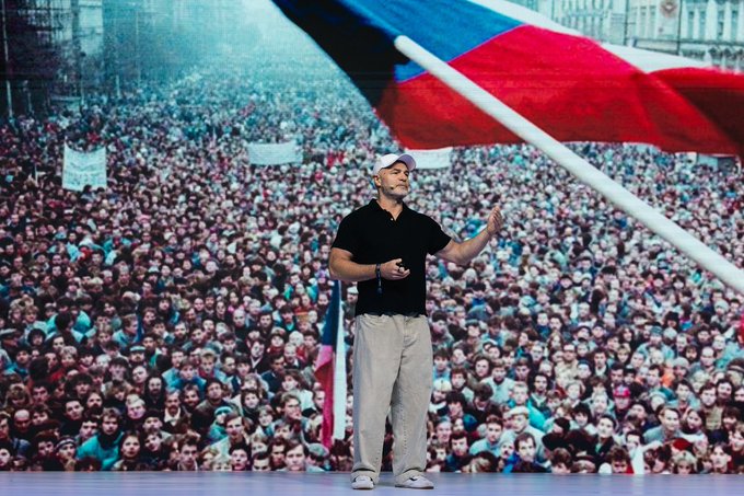 A man in a black t-shirt, beige pants, and white cap stands on stage gesturing with his hand, with a massive projected background showing a huge crowd of people and a flag.