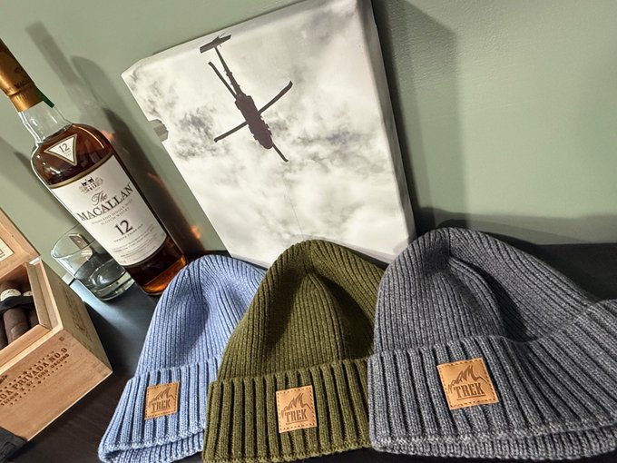 Three knit beanies in blue, olive green, and gray with "TREK" leather patches displayed next to a bottle of The Macallan 12 Year Scotch Whisky and a canvas print of a helicopter.
