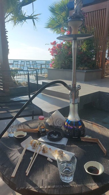A glass hookah sits on a wooden table alongside sushi rolls, chopsticks, dipping sauce, and water glasses at what appears to be a beachside or waterfront lounge setting. The "GM" post likely refers to "Good Morning," with the image showcasing a serene morning hookah session with a scenic view.