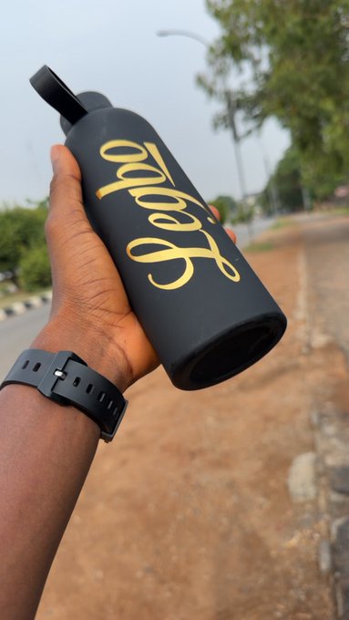 A hand wearing a black fitness band holds a black water bottle with the word 'magic' written in yellow cursive against an outdoor background.