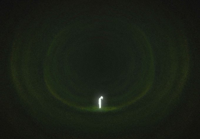 A lone figure in a white suit stands illuminated in the center of a vast, dark void, encircled by a faint, greenish-yellow ring of light.