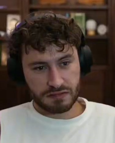 A young man with curly brown hair, a short beard, and a white t-shirt wears large headphones while looking off-camera with a serious, contemplative expression, set against a bookshelf background. The image conveys a focused, thoughtful reaction—befitting the post's reflective tone about OG perpetual futures traders having the last laugh over crypto newcomers.