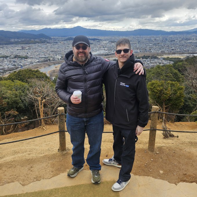 Two men pose together on a scenic overlook in Japan, with one wearing a jacket bearing a "Hoskinson" name patch, sharing a friendly moment with arms around each other against a panoramic backdrop of a sprawling city and distant mountains. The image captures a personal encounter with Charles Hoskinson, the Cardano founder referenced in the post, celebrating his contributions to crypto and his advocacy before Congress.