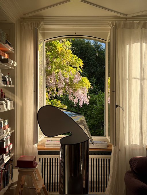 A glossy black sculptural chair or modern art piece sits on a pedestal in front of a large arched window draped with sheer curtains, framing a view of blooming wisteria and green foliage. Bookshelves filled with books line the left side of the cozy, sunlit room.