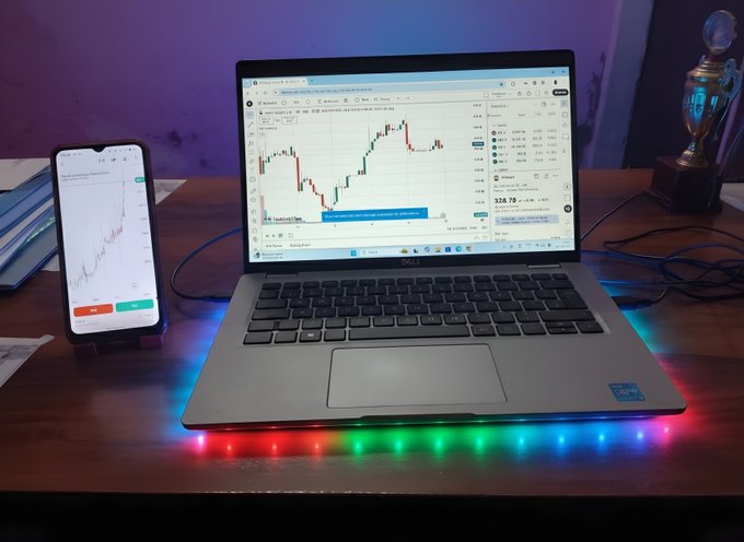 A laptop displaying a cryptocurrency or stock trading chart sits on a desk with colorful RGB LED lights along its base, accompanied by a smartphone showing a similar trading interface on the left and a trophy on the right.