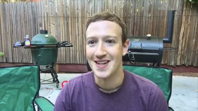 Mark Zuckerberg smiles in a purple t-shirt during what appears to be a video call, with a green Big Green Egg grill and black smoker visible in a backyard setting behind him.