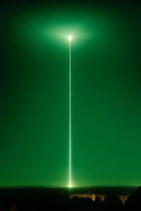 A nighttime photograph of a bright green beam of light shooting vertically into the sky above a distant cityscape, likely a laser or searchlight display.