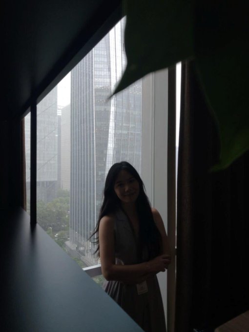 A young woman with long dark hair stands smiling beside a large window overlooking a modern cityscape with skyscrapers and greenery visible below.