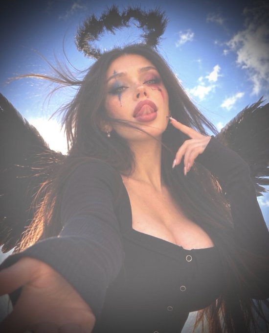 A woman poses for a close-up selfie styled as a dark angel, with black feathered wings and a halo framing her head against a bright, cloudy sky, while smudged makeup and exaggerated lips complete the dramatic look—fitting the playful "$Clussy angel" crypto meme aesthetic of a mischievous, attention-seeking figure.