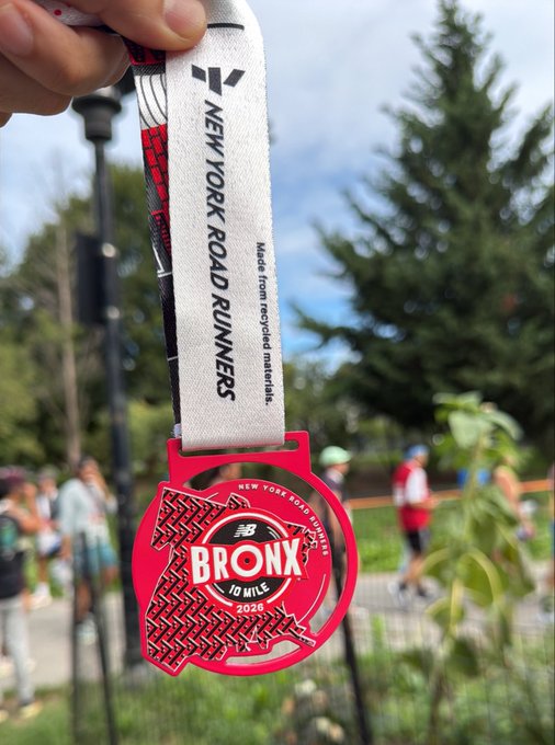 A hand holding a red 'BRONX 10 MILE 2026' New Balance medal from the New York Road Runners, noting it is 'Made from recycled materials.'