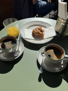 A table set with a croissant on a white plate, a glass of orange juice, and two cups of coffee on saucers with sugar packets, bathed in bright sunlight.