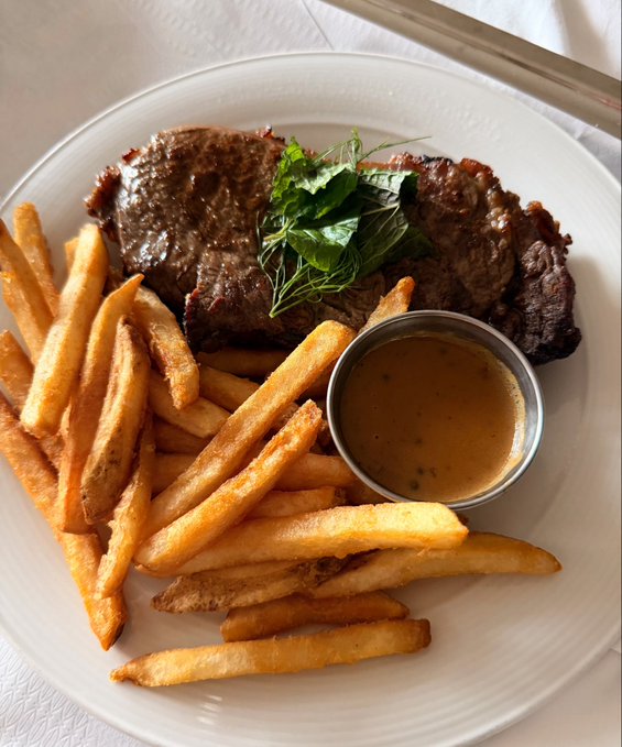 A plate of steak with french fries and a side of brown sauce, garnished with fresh greenery on top of the meat.